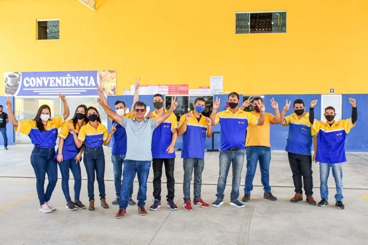 Equipe uniformizada, de camisas azul e amarela, sorrindo e fazendo o sinal de paz diante de uma loja de conveniência.